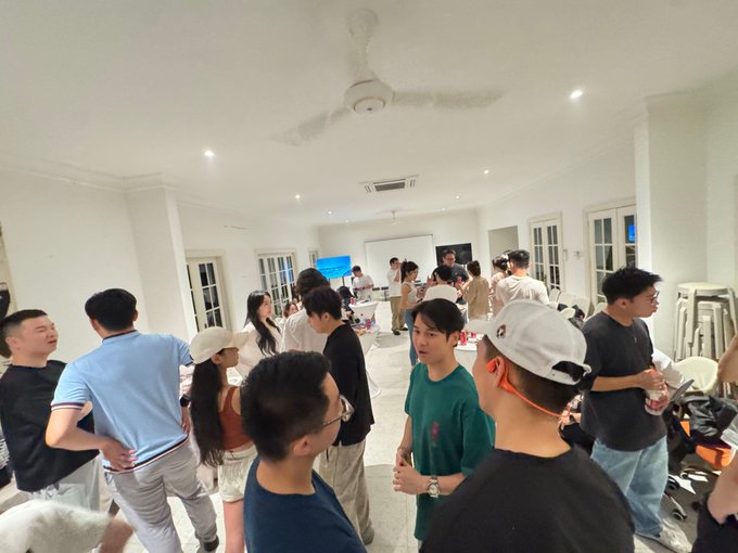 A crowded indoor gathering with many attendees standing and mingling in a white room with a ceiling fan.
