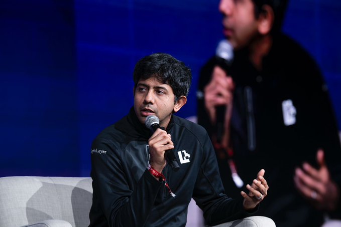 The image features a man sitting down, holding a microphone, possibly giving a speech or presentation. The background shows another person standing behind him, likely an audience member. There are also two more people visible in the scene, one of them wearing a black jacket with white lettering on it and the other dressed in all-black attire.