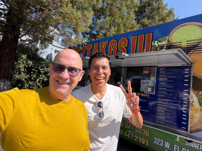 Two men smiling for a selfie in front of a food truck called 'GYROTAKAS III' with visible phone numbers 323-848-0236, 818-841-2191, and an address on 323 W.