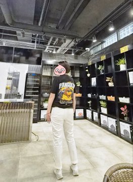 A woman wearing a "Diadora" branded graphic shirt, white pants, and glasses, with her face pixelated, standing in what appears to be a retail store with shelving units displaying various products.