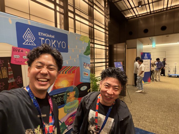 Two smiling attendees pose for a selfie in front of the "ETHGlobal TOKYO" event banner, wearing event lanyards inside a conference hall. Other participants can be seen mingling in the background as they enjoy the Ethereum-focused hackathon gathering.
