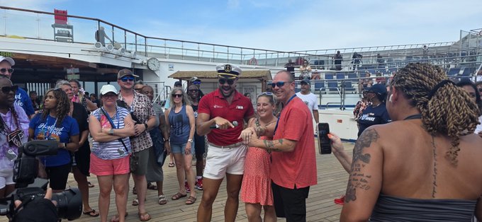 A group photo taken on the deck of a cruise ship, featuring a smiling captain in uniform posing with several passengers in red shirts while surrounded by other tourists.