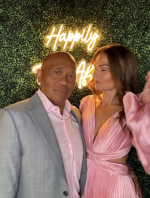 A couple poses together in front of a neon "Happily" sign, with the woman in a pink pleated dress looking affectionately at her partner in a light blue suit and pink shirt. The image pairs the romantic backdrop with the post's reflection on a season of major life transitions—selling their home, moving, and celebrating milestones—capturing a moment of calm amid the chaos.