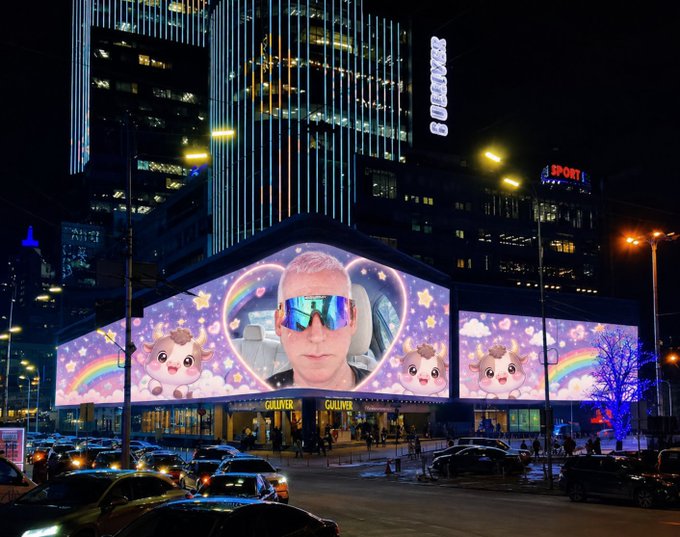 A massive LED billboard outside the Galiver shopping center displays a surreal animated scene featuring a man wearing reflective rainbow sunglasses surrounded by cute cartoon calves, rainbows, and stars, with the name "GALIVER" visible on the storefront and "SPORT" and "Unilever" signage on surrounding buildings. The nighttime cityscape, filled with traffic and pedestrians, highlights how the image matches the post's description of launching a $JAMES crypto animation advertisement on a prominent shopping center screen.
