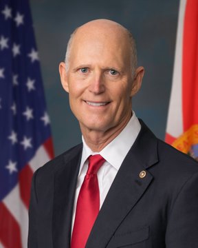 An official portrait of a bald man in a dark suit with a red tie, smiling in front of an American flag and a Florida state flag.
