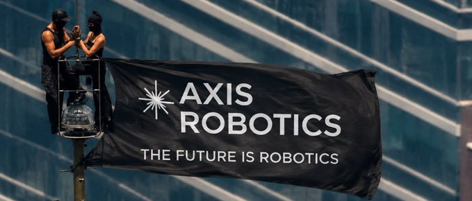 Two people in a bucket lift install a black banner reading 'AXIS ROBOTICS - THE FUTURE IS ROBOTICS' against a glass building facade.