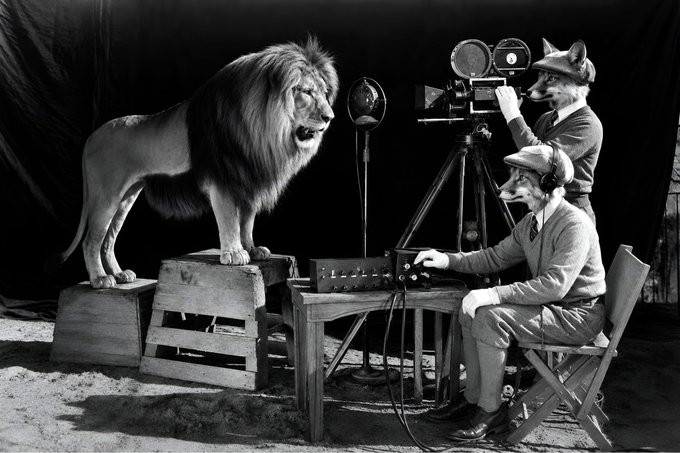 A vintage black-and-white photograph shows MGM's famous lion roaring into a vintage microphone, with two men wearing lion masks operating a film camera and sound equipment.