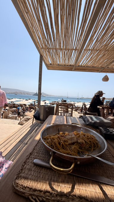 A plate of spaghetti and meat sits on a table at a beachside restaurant under a bamboo canopy, with the ocean and lounge chairs visible in the background.