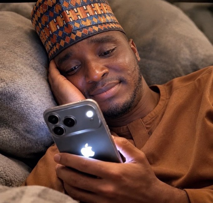 A man wearing a patterned cap and brown shirt smiles softly while looking at his iPhone, lying down against a gray pillow.