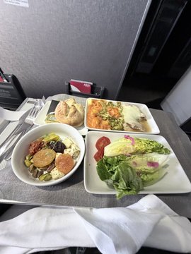 An airline meal tray with a bread roll, a small salad with olives, a main dish with vegetables, and a green salad with dressing.