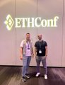 The image features two men standing next to a large white sign that says "ETHCONF" written in black letters. The sign is illuminated, making it stand out against the background. Both men are dressed casually, with one wearing a white shirt and the other wearing a black shirt. They appear to be posing for a photo together, possibly at an event related to cryptocurrency or blockchain technology.