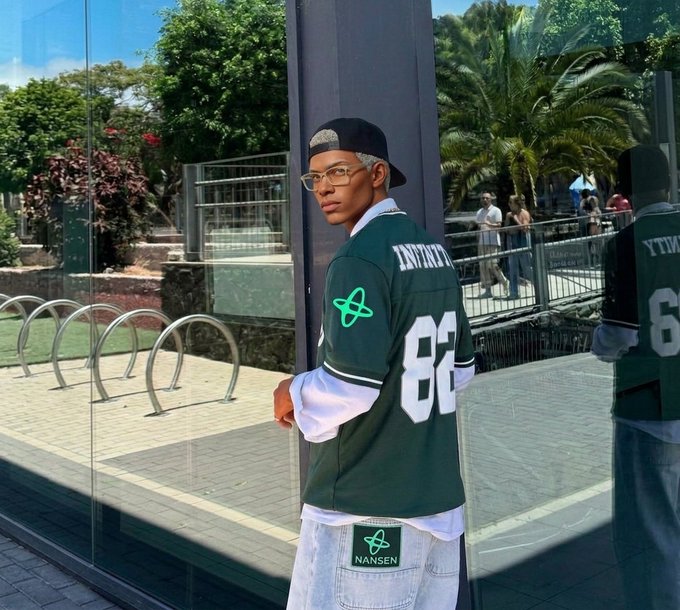 A young man wearing a green jersey with 'INFINITY 82' on the back and a 'NANSEN' logo stands in front of a glass window, looking over his shoulder.