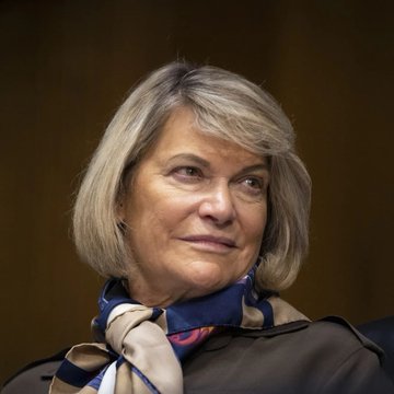 The image shows a woman with shoulder-length gray hair, wearing a dark blazer and a colorful silk scarf in blue and beige tones, looking off to the side with a subtle smile against a blurred brown background. The accompanying post identifies her as Senator Lummis, pictured here as she advocates for the Clarity Act to protect crypto exchange customers during bankruptcies, ensuring their assets are treated like those in traditional finance.