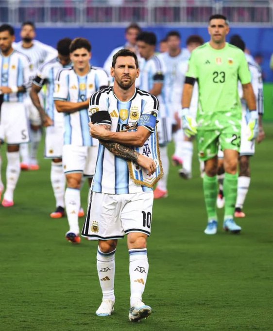 Lionel Messi wearing the Argentina national team jersey (number 10) with captain's armband, walking on a soccer field with teammates including goalkeeper wearing number 23.