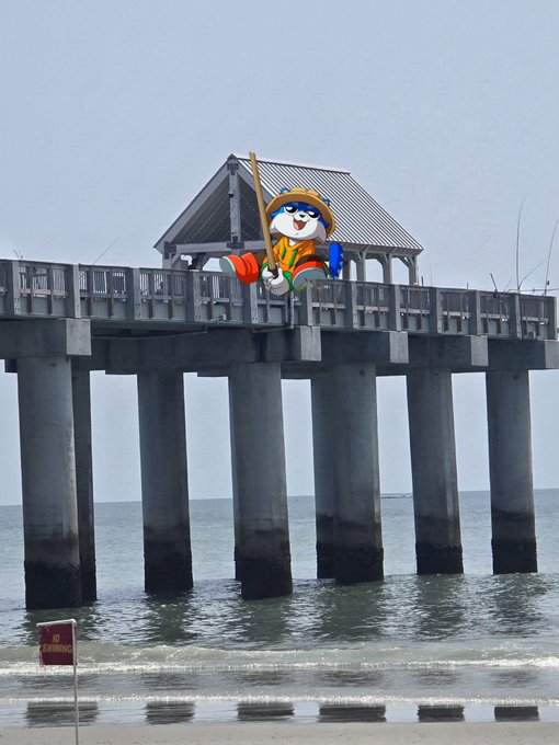Photograph of a pier extending over the ocean with a cartoon cat mascot in a straw hat, orange shorts, and blue shirt sitting on the railing holding a fishing pole.