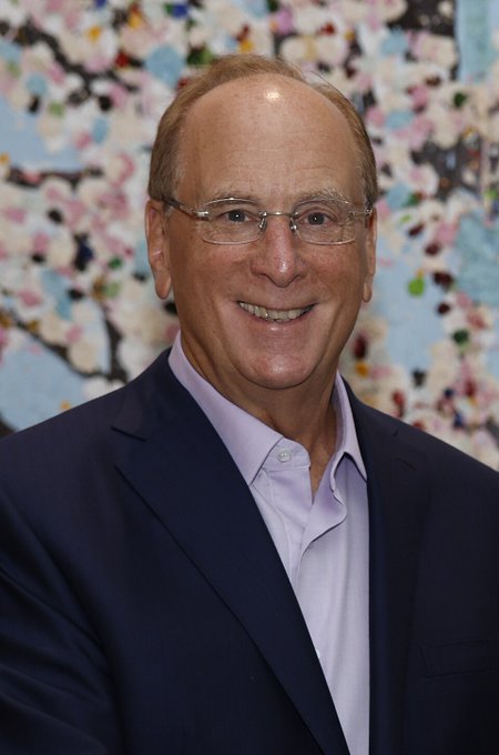 A portrait photograph shows a smiling, older man with a receding hairline, wearing rimless glasses, a navy blue suit jacket, and a light purple button-down shirt, posing against a backdrop depicting pink cherry blossoms.