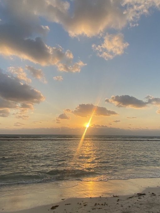 A golden sunrise over the ocean, with the sun peeking through scattered clouds and casting a shimmering reflection across the gentle waves onto the sandy beach. The serene "gm legends" morning vibes capture the peaceful start of a new day.