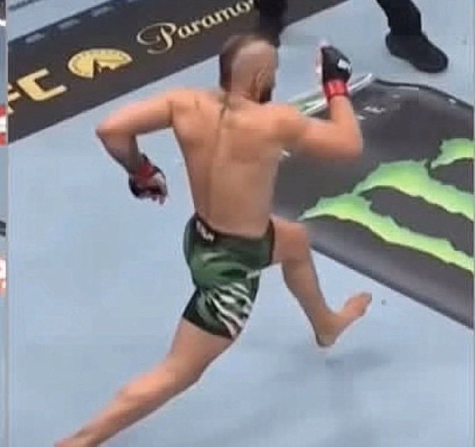 Action shot of a shirtless MMA fighter in camouflage shorts celebrating or running inside an octagon with Monster Energy and Paramount branding visible in the background.