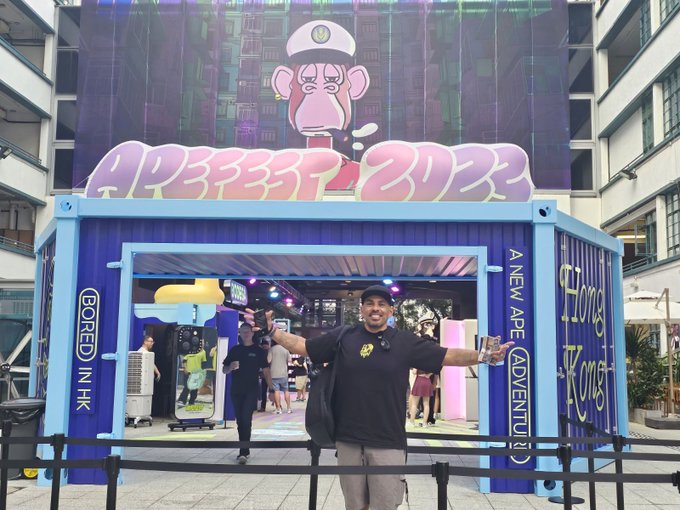 A man wearing a black shirt stands next to a blue building with a large poster featuring a cartoon monkey, which reads "Aprefest 2020". The man appears to be posing for a photo or selfie near the entrance of this building.