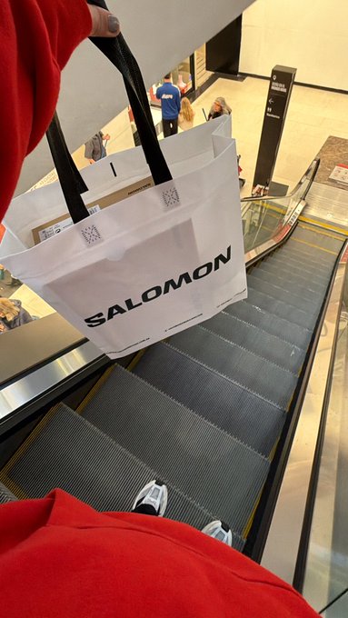 A first-person perspective photo looking down at a Salomon shopping bag held while riding an escalator, with the rider wearing red pants and white sneakers.