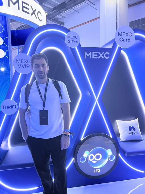 A man wearing a white t-shirt and lanyard stands at a brightly lit MEXC cryptocurrency exchange booth featuring neon blue lighting and signs reading 'MEXC 0-Fee', 'MEXC Card', 'MEXC VVIP', and 'TradFi'.