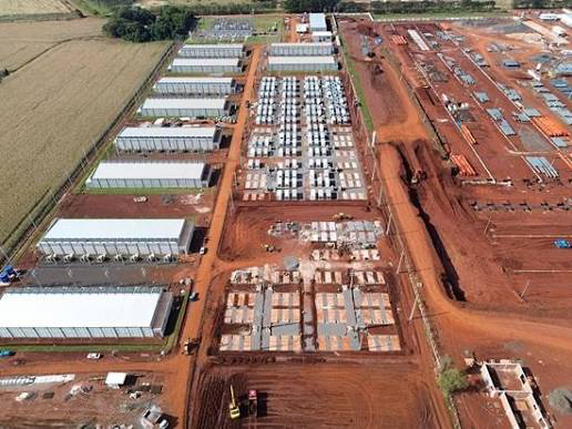 An aerial view shows rows of industrial buildings and containerized structures at a large-scale facility, with exposed red dirt indicating ongoing construction and expansion of data center or mining infrastructure. The image relates to HIVE Digital Technologies by visually depicting the type of operational footprint—a growing crypto mining or data center site—whose Swedish VAT assessments and accounting disclosures delayed the company's quarterly financial filing.