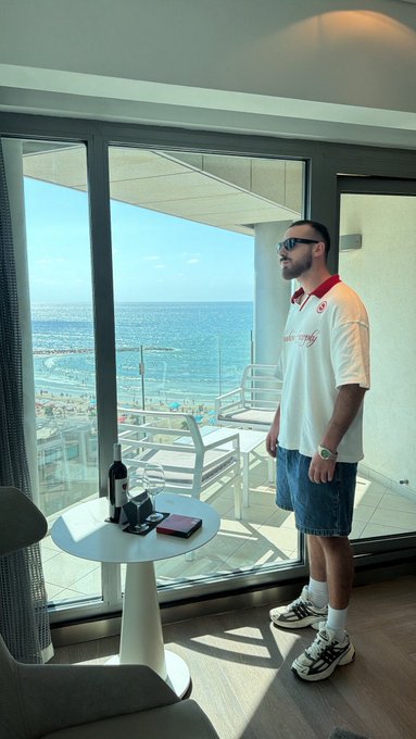 A man in sunglasses, a white polo shirt, and denim shorts stands on a balcony overlooking the ocean and beach on a sunny day, with a small table holding a wine bottle nearby, capturing the relaxed morning vibes of the "Gm friends" greeting.