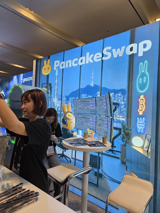 Two women pose for a selfie inside a PancakeSwap-branded booth at an event, with the company's logo, crypto symbols, and a city skyline backdrop visible behind them.