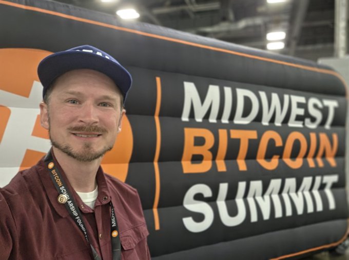 A man wearing a blue cap and maroon shirt takes a selfie in front of an inflatable 'MIDWEST BITCOIN SUMMIT' sign, wearing a lanyard.