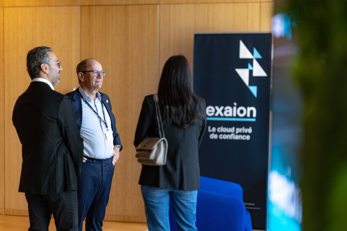Two men in suits and a woman with a beige handbag stand conversing in front of a banner for 'exaion' with the tagline 'Le cloud privé de confiance'.