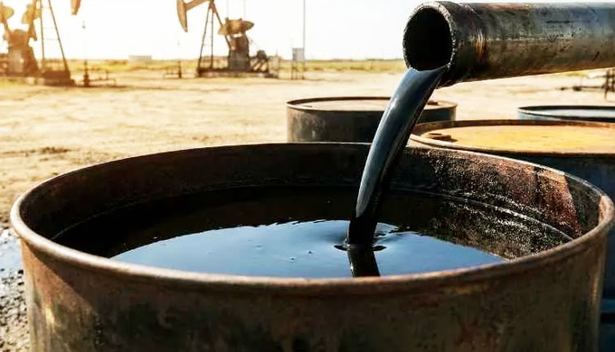 Crude oil being poured from a pipe into a metal barrel in an oil field setting with pumpjacks visible in the background.