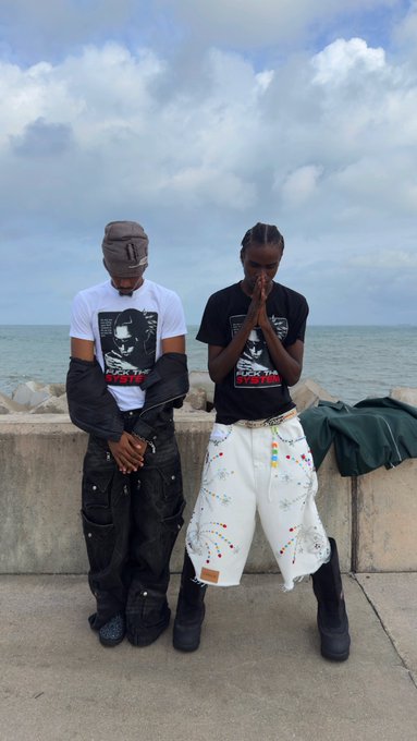 Two people pose by the seaside, both wearing matching F*CK THEM graphic tees, with the left person in a balaclava and dark cargo pants while the right person stands in prayer pose wearing white embellished shorts, embodying the post's "good morning, it's Monday, let's print!" mantra with their bold, ready-to-conquer-the-week energy.