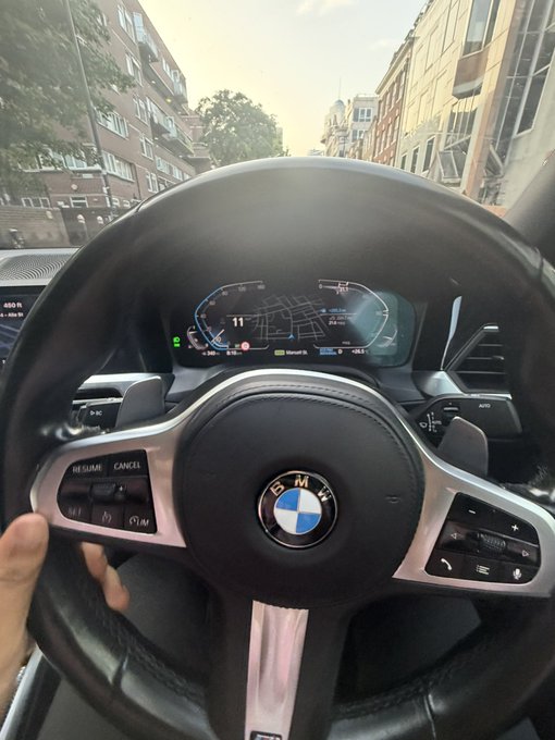 A driver's perspective inside a BMW shows the steering wheel with the iconic blue and white logo and a digital dashboard displaying a speed of "11," all captured with the playful caption "GM bros" greeting fellow car enthusiasts.