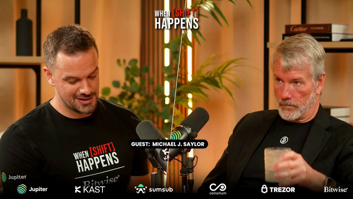 The image features two men sitting next to each other with microphones in front of them, suggesting they are hosting an interview or podcast discussion. The background includes a bookshelf filled with various items such as bottles and books, indicating that this might be taking place in a living room or office setting. The post contains text discussing Bitcoin's dominance and the importance of protocols, which is related to the conversation between the two men on the podcast.