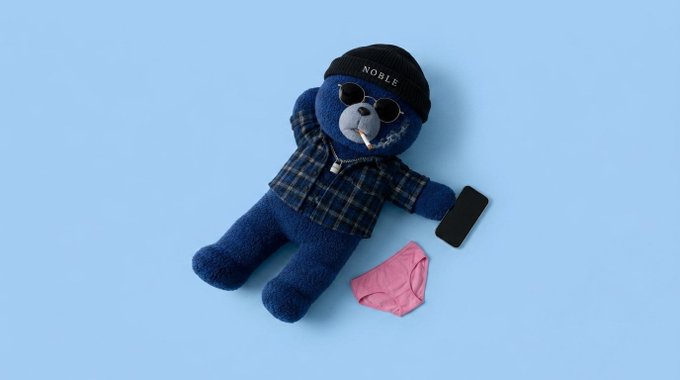 A blue teddy bear wearing sunglasses, a beanie labeled 'NOBLE', a plaid shirt, and a gold chain lies on a light blue background next to a smartphone and pink underwear.