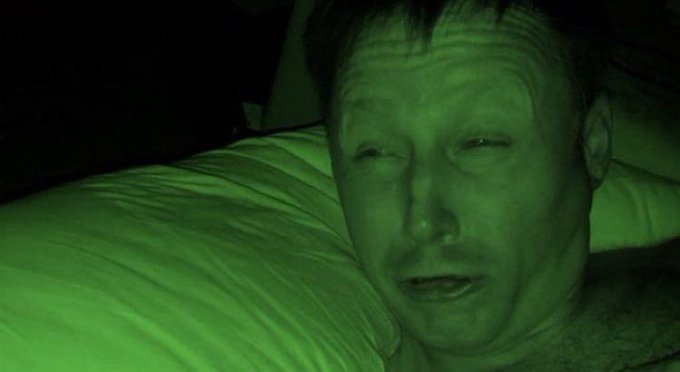 A green-tinted night vision close-up of a man's face looking tired or distressed as he lies against a white pillow.