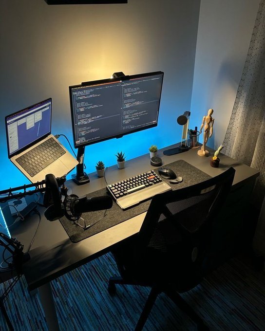 A modern home office desk setup featuring a laptop and curved monitor displaying code, illuminated with blue LED backlighting, accompanied by a microphone, keyboard, and small desk plants.