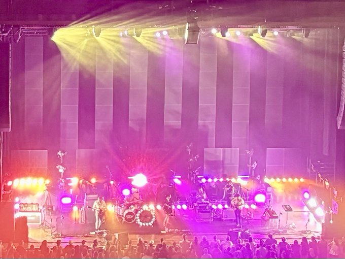 A concert stage is illuminated with intense pink and yellow lights, showing musicians performing behind instruments and equipment.