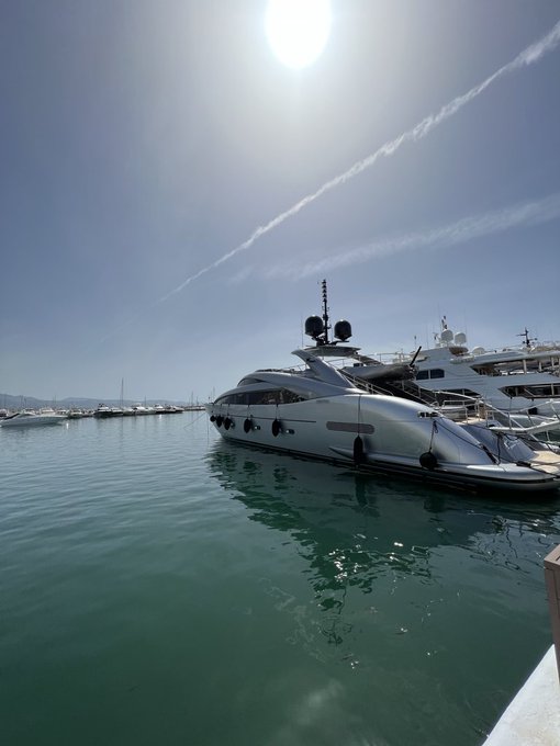 A sleek white luxury yacht docked in a sunny marina with turquoise water, other boats in the background, and a clear blue sky with a jet contrail.