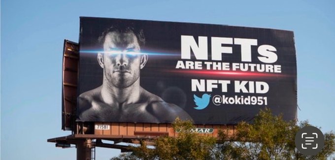 A billboard against a blue sky displays a black-and-white photo of a muscular man with glowing blue eyes, accompanied by the text "NFTS ARE THE FUTURE" and "NFT KID" with the Twitter handle "@kokid951". The post notes this is a five-year-old advertisement (from 2021) that the poster claims is still relevant today.
