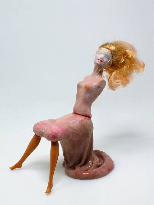 A surreal sculpture of a seated female figure with blonde flowing hair, an elongated neck, and wearing a long pink dress, displayed against a plain white background.