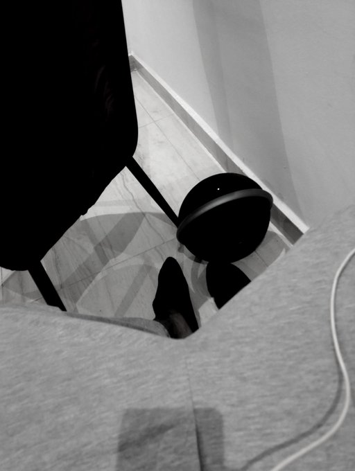 A black-and-white photo showing a reflection on a floor, featuring a dark chair and a person's feet next to a cylindrical object.