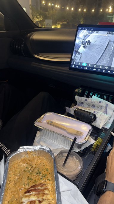 Inside a car, a passenger's meal is spread on their lap including a foil container of crepes or pancakes, a wooden cutlery set, a drink, and other snacks, while a tablet screen displays an exterior camera view.
