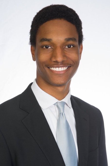 A professional headshot of a smiling young Black man wearing a dark suit, white shirt, and light blue tie against a white background.