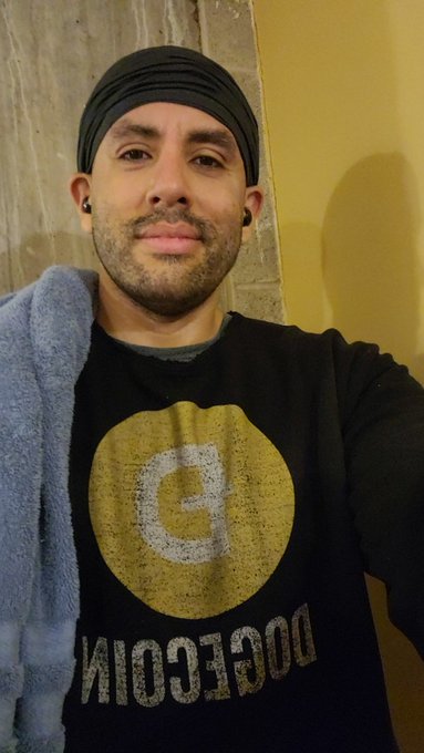 The image features a man wearing a black sweatshirt with a yellow circle design that says "dogecin" on it, holding a towel over his head as he takes a selfie. The caption of the photo includes a reference to a quote from the year 2002 and mentions that the man is a dogeoin.