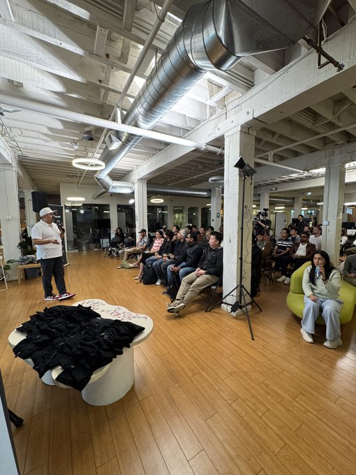 A man with a microphone speaks to a seated audience in an industrial-style venue with exposed ceiling ducts, while a camera on a tripod records the event.