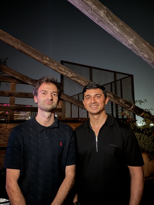 A nighttime photograph of two men smiling at the camera, one wearing a black polo shirt and the other a dark zip-up shirt, standing in front of a wooden structure with metal mesh.
