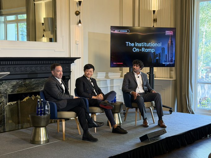 Three men in business attire sit on stage in chairs during a panel discussion titled 'The Institutional On-Ramp,' with the screen displaying 'NEXT SESSION' and 'TOKYO SUMMIT' alongside imagery of Tokyo City.