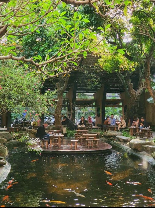 An outdoor café or restaurant with seating areas surrounded by lush trees and a large pond filled with koi fish.