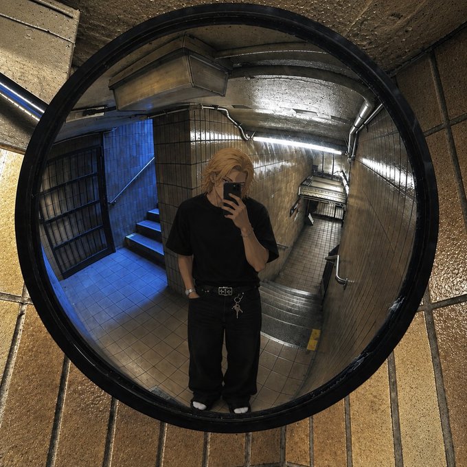 A mirror selfie of a person with blonde hair dressed in all black with keys hanging from a belt, standing in a tiled stairwell illuminated by blue and white lighting.